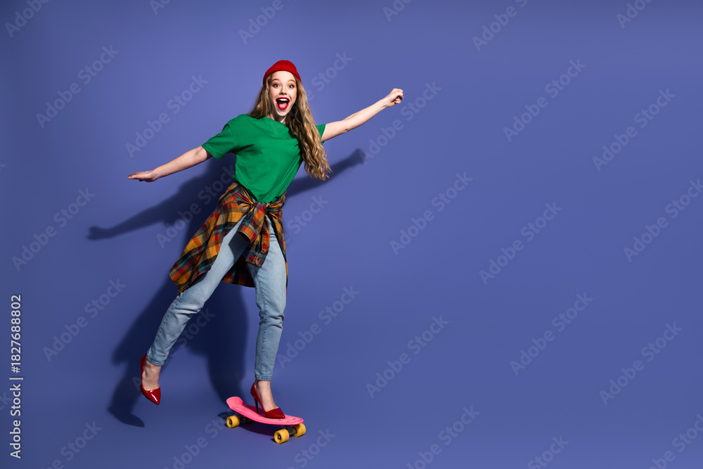 Fototapeta premium Young girl on a skateboard in a green shirt red beanie plaid shirt jeans and red heels smiling against a blue background casual stylish lifestyle fashion shot
