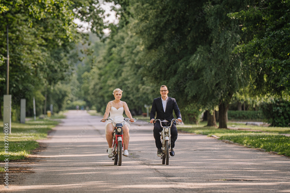 Fototapeta premium Couple riding bicycles on a tree-lined path, showcasing joy and connection, dressed in formal and casual attire, capturing a moment of togetherness and celebration