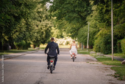 Wallpaper Mural Man in formal suit and woman in wedding dress riding bicycles on a tree-lined street, capturing a joyful moment of celebration and love in a serene outdoor setting Torontodigital.ca