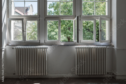 An interior perspective of a row of modern white-framed windows, several of which are tilted open for ventilation. The view outside reveals lush green trees, a blue sky with clouds.