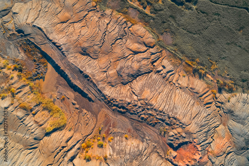 Abstract aerial view of sculpted, eroded industrial waste (tailings). The terracotta red earth shows intense geological flow patterns, sharply contrasting with the dark green natural boundary.