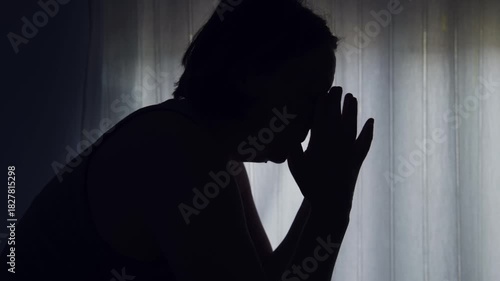 Silhouette of sad woman sitting by window with head in hands, feeling depressed and lonely at home. 4K.