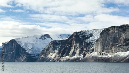 Breathtaking cliffs rise majestically above calm waters on a summer day. The clear sky showcases the beauty of the Arctic landscape with snow-capped peaks.