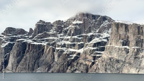 Breathtaking cliffs rise majestically above calm waters on a summer day. The clear sky showcases the beauty of the Arctic landscape with snow-capped peaks.