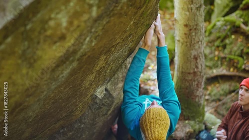 slow motion rock climbing outdoors in autumn forest. friends rock climbers climbs bouldering belaying each other. man in bright blue hoodie and yellow knitted cap hanging on edge of overhanging cliff