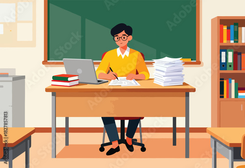 A teacher in a classroom sits at his desk, working on paper with a laptop and books