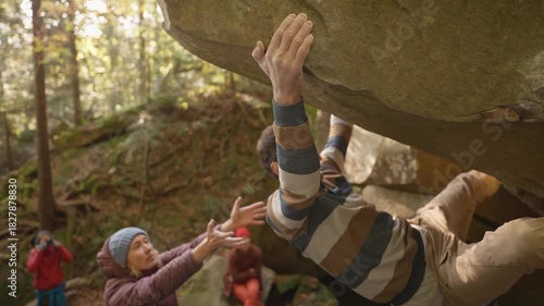 man rock climber climbs on overhanging boulder by it edges using wide hands grips, friends belay him. climbing outdoor in autumn forest on natural rocks. healthy active lifestyle, training outdoors.