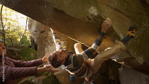 man rock climber climbs on overhanging boulder by it edges using wide hands grips, friends belay him. climbing outdoor in autumn forest on natural rocks. healthy active lifestyle, training outdoors.