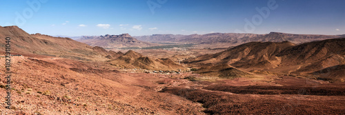 A wide view shows red rocky mountains and valleys in Morocco. In the distance, a small town nestles between the mountains under a bright, clear blue sky