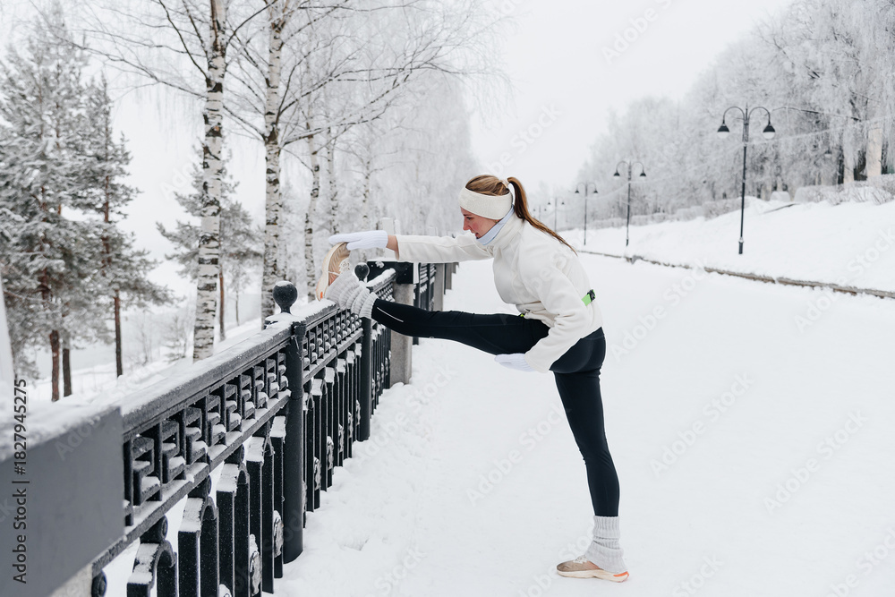 Naklejka premium young caucasian woman doing stretching exercises before running in park in winter, white sports jacket, black trousers, active lifestyle concept, jogging in winter, sports life, copy space