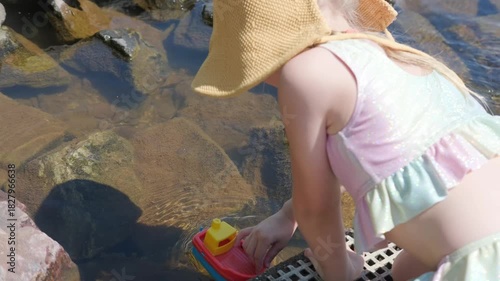 girl in swimsuit and sun hat playing by river with plastic toy boat, red sail floats on surface water on sunny day play and childhood, enjoying warm sunny day and carefree summer outdoor fun