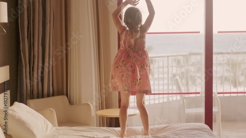 little girl in floral dress jumping on hotel bed, silhouetted against large window with ocean view, arms raised in pure joy and excitement, vacation fun, childhood joy captured in moment carefree play