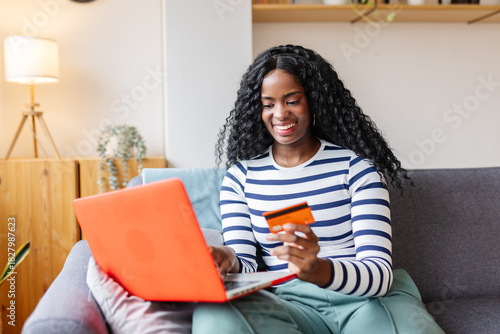 Fotografie Happy Latin American woman using laptop paying online by credit card sitting on sofa