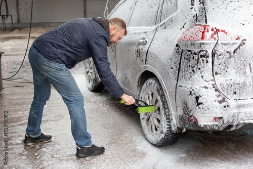 Man scrubbing the wheel of a foam-covered SUV at a self-service car wash. Using green brush, he cleans the rim thoroughly on wet day. Practical auto care, detailing, and seasonal vehicle maintenance.