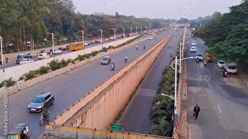 A wide view of a clean urban road with parallel lanes, light traffic, and greenery on both sides. The scene captures early morning or evening city movement with vehicles, trees, and well-maintained