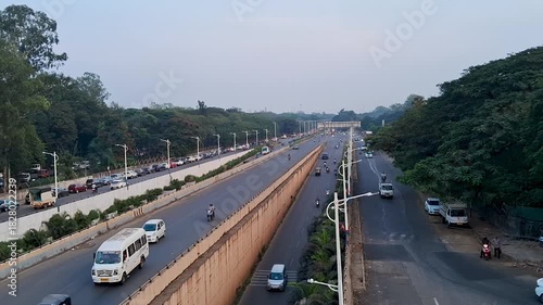 A wide view of a clean urban road with parallel lanes, light traffic, and greenery on both sides. The scene captures early morning or evening city movement with vehicles, trees, and well-maintained