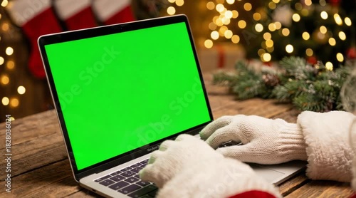 Santa-gloved hands typing on laptop with green screen in festive Christmas setting and warm holiday lights  
