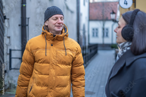 Man wearing orange puffer jacket smiles while talking to a woman in a black coat with ear muffs on a cobblestone street in a charming urban setting with buildings