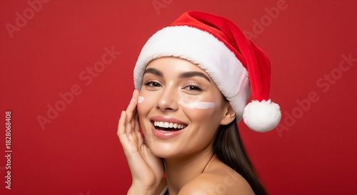 Radiant woman in festive Santa hat smiles brightly, applying nourishing skincare cream to her cheek for a glowing and healthy complexion, embracing holiday beauty and self-care