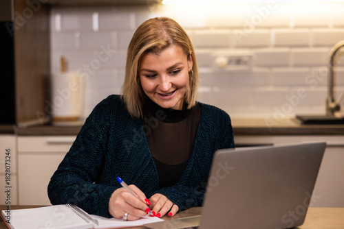 Young woman works on a laptop while taking notes in a modern kitchen setting during the day