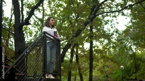 Young woman standing on a cafe veranda with a coffee cup, park backdrop. K