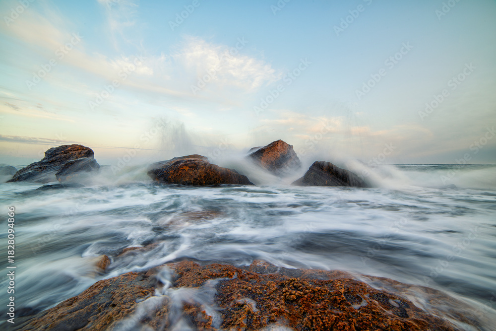Fototapeta premium Long-exposure seascape capturing waves swirling around coastal rocks. The soft motion of water and pastel sky create a serene and powerful marine landscape perfect for nature, travel, and relaxation t