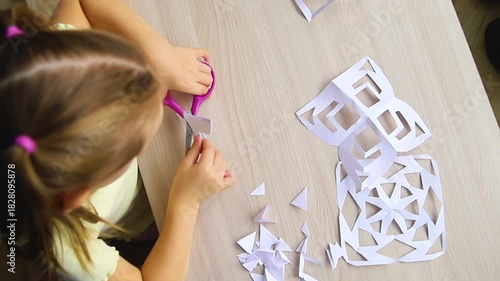 A child creating paper white snowflakes at a table at home. A child cuts patterns on paper with scissors. Decoration for the winter holiday.
