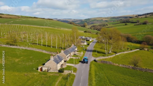 Aerial drone view of a blue tractor driving on a rural road through rolling green hills and