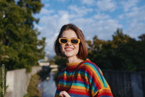 Woman smiling in orange sunglasses and rainbow sweater, portrait outdoors with blue sky, warm sunlight and shoulder bag; happy young adult in colorful casual street style, candid cheerful vibe.