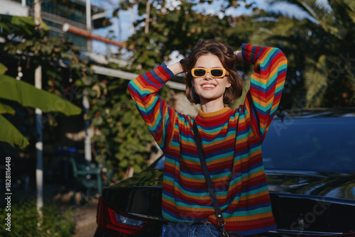 Fotografie Woman in her mid 20s wearing a colorful striped sweater and yellow sunglasses, smiling and leaning against a black car in a tropical outdoor setting, casual outfit with relaxed travel vibe