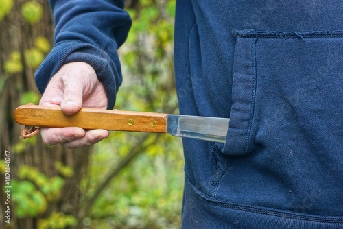 An aggressive man takes out a new, dangerous, sharp, shiny kitchen knife with a wooden handle from the front pocket of his blue raglan jacket during the day on the street.