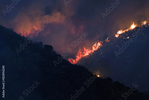 Wildfire raging across a dark mountain slope at night with glowing embers and smoke plumes