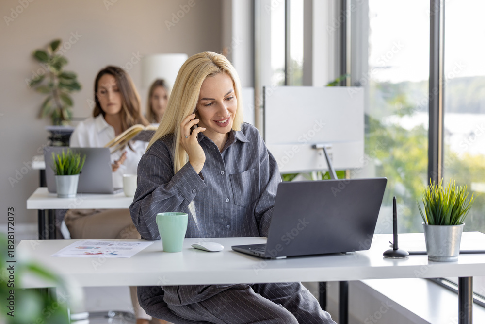 Obraz premium Woman working on a laptop and talking on the phone in a modern office setting with plants and natural light
