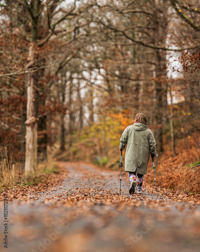 Elderly woman walking alone with trekking poles on a forest path in autumn.