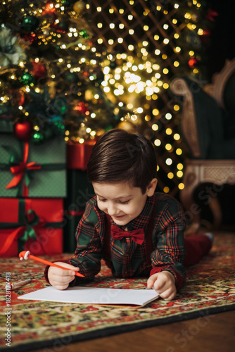A child in a plaid shirt and bow tie writes a letter to Santa under the twinkling lights of a Christmas tree. Pure holiday magic 