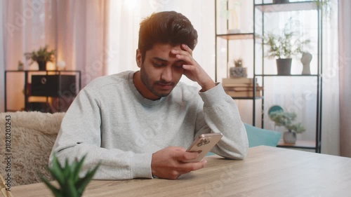 Young Indian man sitting on sofa at table, actively texting on smartphone, thoughtfully resting head on hand in living room at home. Arabian guy looks focused, deeply engaged in conversation or task