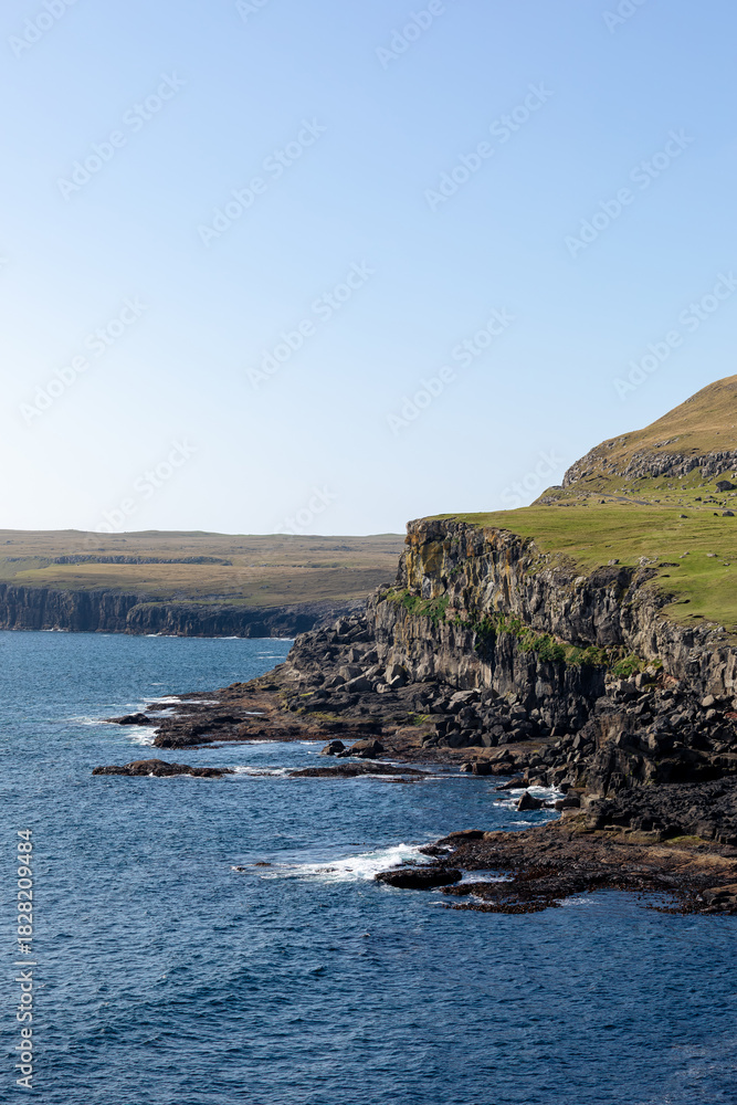 Naklejka premium Panoramic view of the dramatic coastline of the Faroe Islands, featuring steep volcanic cliffs, deep-blue North Atlantic waters and wide open green hills under a clear sky.