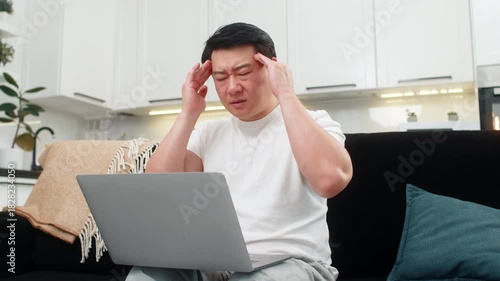 Asian man at home works on laptop feels headache rubs temples closes eyes a moment and breathes slow. Chinese guy on sofa puts laptop aside takes pause does deep breath feels relief and rests quiet