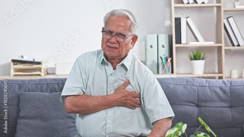 Asian Elderly man holding chest pain couch showing concern about health care and heart symptom while seated home senior male clutching chest sofa experiencing discomfort living room showing concern