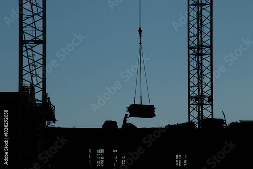Blue-Black Silhouette of Worker in Hard Hat directing Cargo on Construction Site surrounded by Construction Machinery and Materials. Hook of Tower Crane lifts Load next to Builder.
