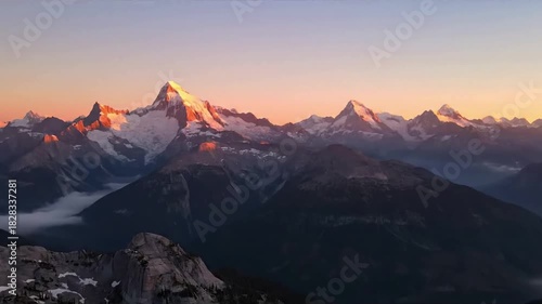 Majestic, snow capped mountain range at sunrise, with deep valleys filled with ethereal mist and golden light illuminating rugged peaks. Wide aerial shot slowly panning across the vast wilderness.