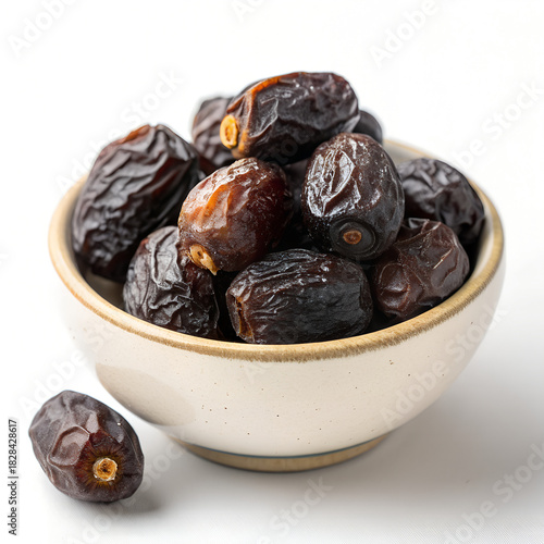A photo of dark, wrinkled dates piled in a small, cream-colored bowl on a white surface, softly lit, creating a simple, inviting still life.