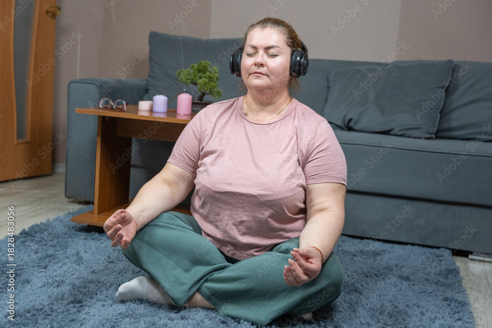 Fototapeta premium Plus-size woman practicing mindfulness meditation in living room, seated cross-legged on soft rug wearing headphones, comfortable casual clothing, serene atmosphere with candles and houseplant