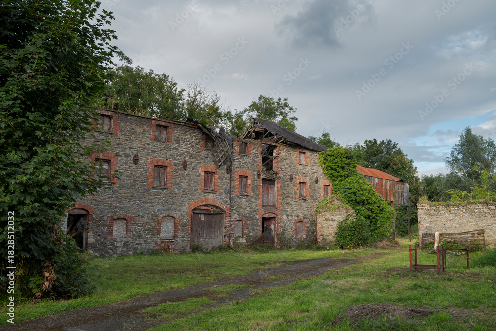 Fototapeta premium Ruins of an abandoned, industrial building shows crumbling stone and brick, with boarded-up windows and ivy climbing the surface. Photo taken in Inniskeen, County Monaghan, Ireland