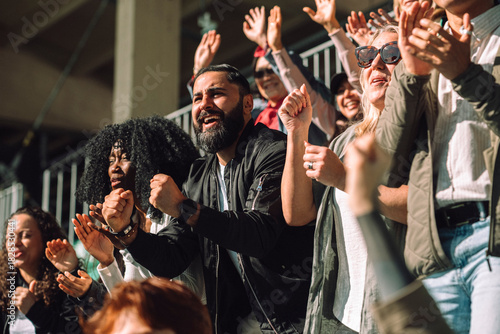 Happy enthusiast man making fist while cheering from sports stadium stand with other audience