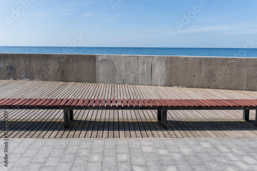 A view of the calm Baltic Sea from an empty embankment. The horizon and blue sky are visible. A wooden bench and a gray plastered parapet are visible. The background is a landscape.