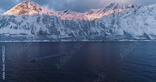 Cinematic aerial drone footage of a small fishing boat leaving a wake across dark Arctic fjord below dramatic snow-covered mountains at golden sunrise in Senja, Northern Norway.