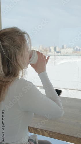 The caucasian young woman is drinking coffee in a coffee shop in winter in front of a window