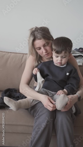 The mother is putting socks on her child while sitting on the sofa at home 
