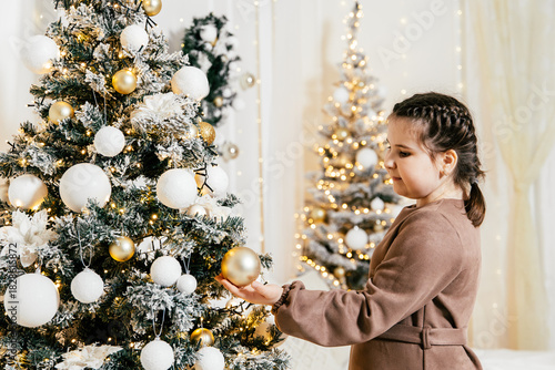 Child decorates Christmas tree with golden ornaments and white decorations in festive indoor setting